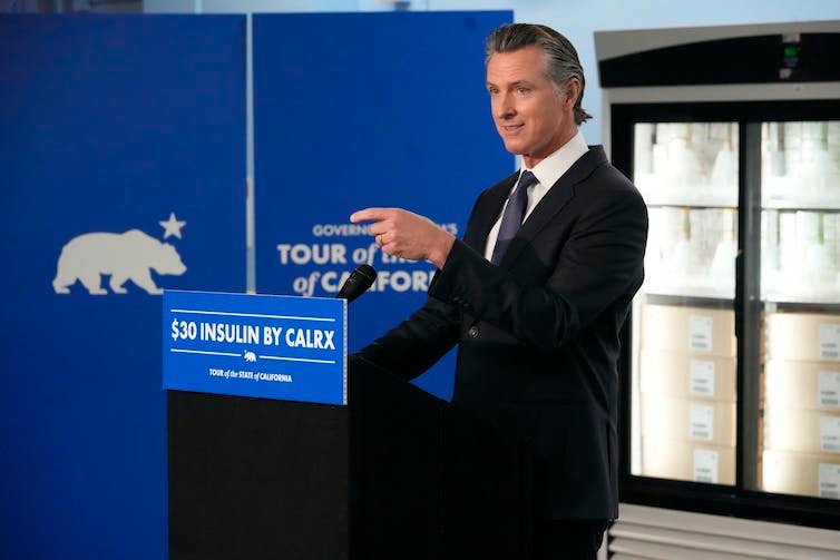 A man in a business suit points in front of a lectern that says $30 worth of insulin, with insulin coolers in the background.