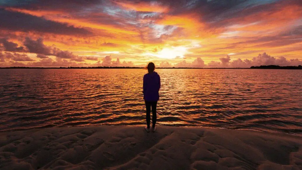 A woman watches the sunset with her back to the camera in front of a body of water