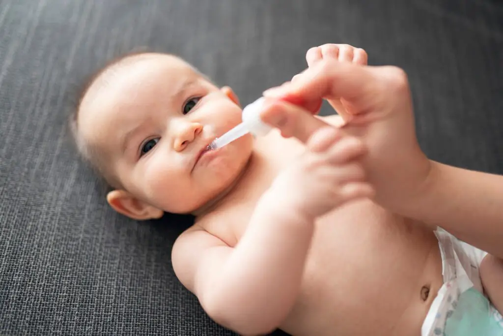 The child receives vitamins through a dropper from an adult hand.