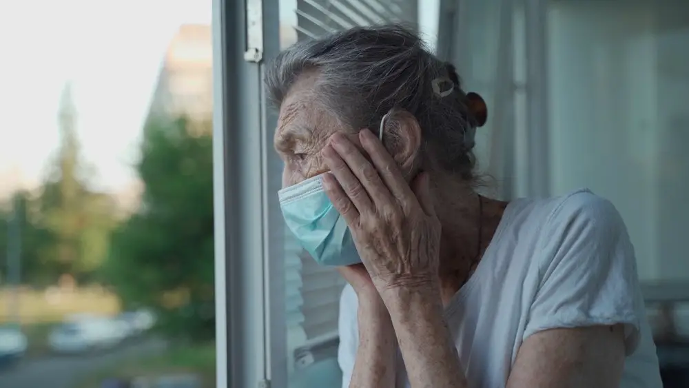 The elderly woman in a medical mask looks out the window with concern